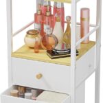 White and wood rolling makeup organizer cart with divided top, open shelf, and two fabric drawers, filled with cosmetics.